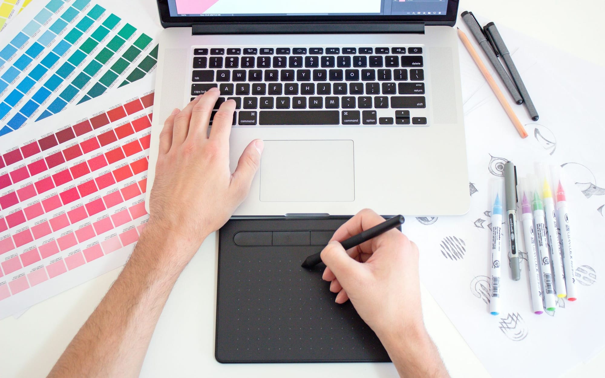 Hände an Laptop und Grafiktablett, daneben Farbfächer und Stifte – Design und Code aus einer Hand