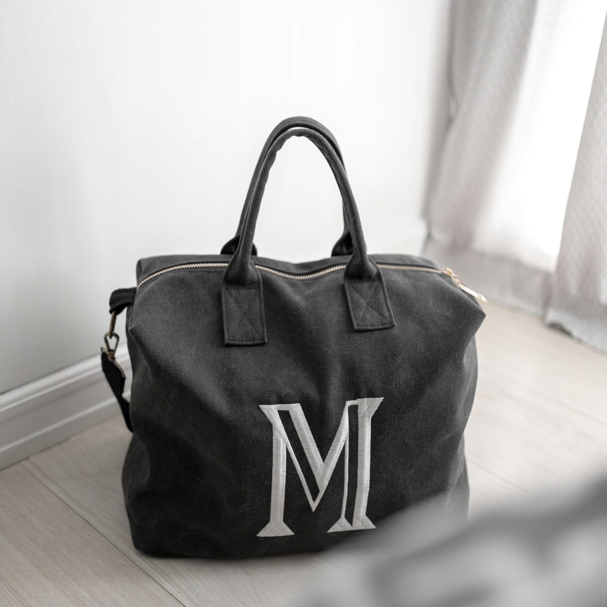 Dunkelgraue Canvas-Tasche mit großem, weiß gesticktem Monogramm „M“ auf hellem Holzboden vor weißem Vorhang